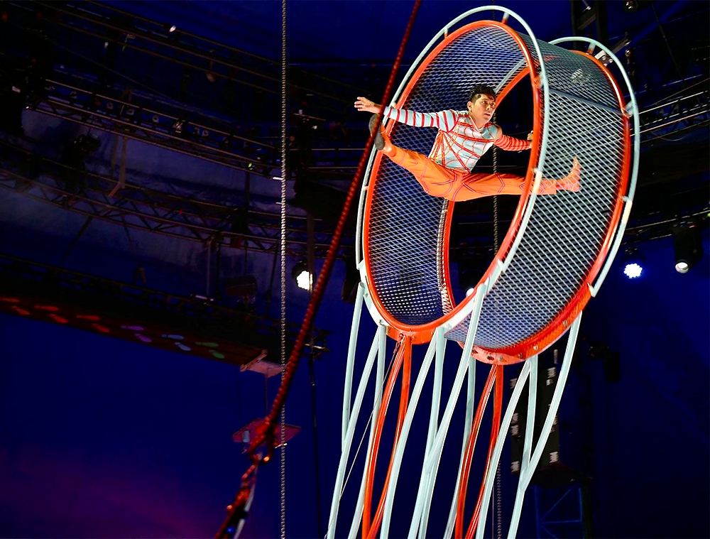 Big Apple Circus Hometown Playground: an aerialist in a red and silver striped costume with orange tights does a split inside a spinning wheel. Costume design by Cynthia Nordstrom