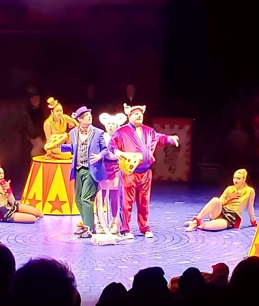 Big Apple Circus Hometown Playground: clowns in a purple top hat and in white and red mouse costumes carry a giant wedge of cheese past performers in gold sequins. Costume design by Cynthia Nordstrom