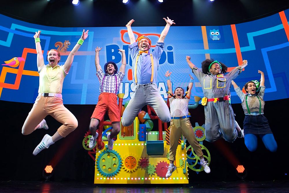 Blippi and cast in bright overalls and caps jumping on stage, Blippi The Musical. Costume design by Cynthia Nordstrom