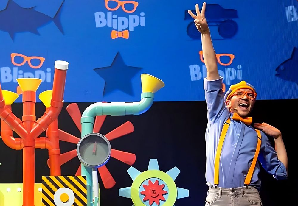 Blippi in a blue shirt and orange bow tie waving before a pipe and gear set, Blippi The Musical. Costume design by Cynthia Nordstrom