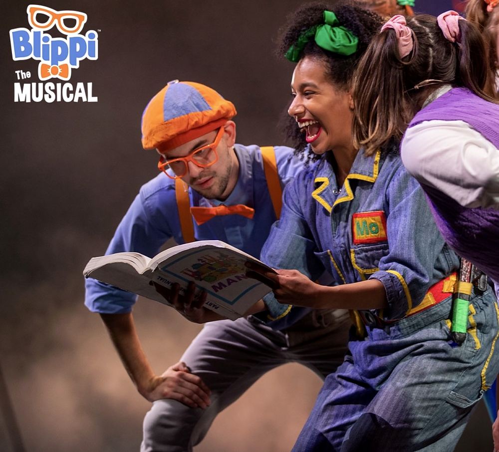 Blippi in his orange cap and glasses reading with Meekah in gray overalls, Blippi The Musical. Costume design by Cynthia Nordstrom
