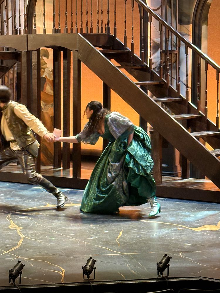 Young man in tan running with a woman in a green gown by a staircase, The Three Musketeers. Costume design by Cynthia Nordstrom