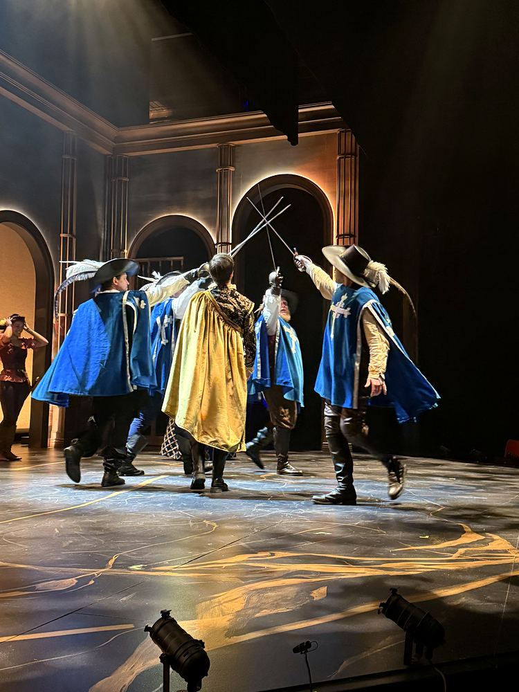 Musketeers in blue and gold capes dueling with swords, The Three Musketeers. Costume design by Cynthia Nordstrom