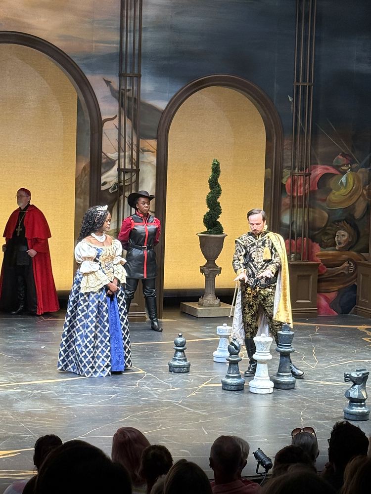 Courtiers in a blue patterned gown and a gold brocade coat by a giant chess set, The Three Musketeers. Costume design by Cynthia Nordstrom