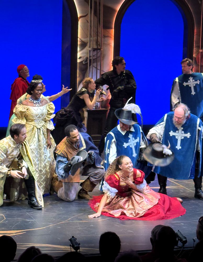 Musketeers in blue tabards and a lady in gold around a woman in a red gown, The Three Musketeers. Costume design by Cynthia Nordstrom