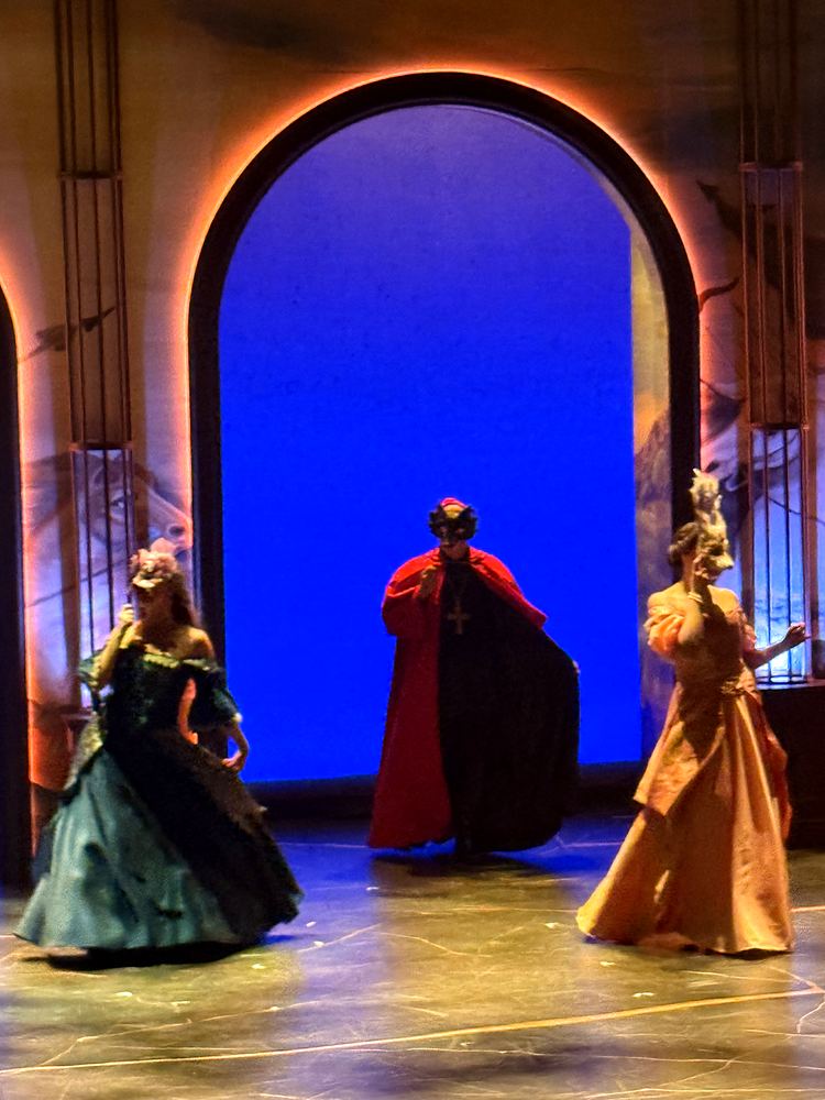 Two women in green and gold gowns flanking a cloaked figure in red, The Three Musketeers. Costume design by Cynthia Nordstrom