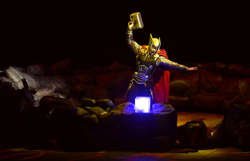 Thor in gold armor raising his hammer over a glowing blue cube, Marvel Universe Live. Costume design by Cynthia Nordstrom