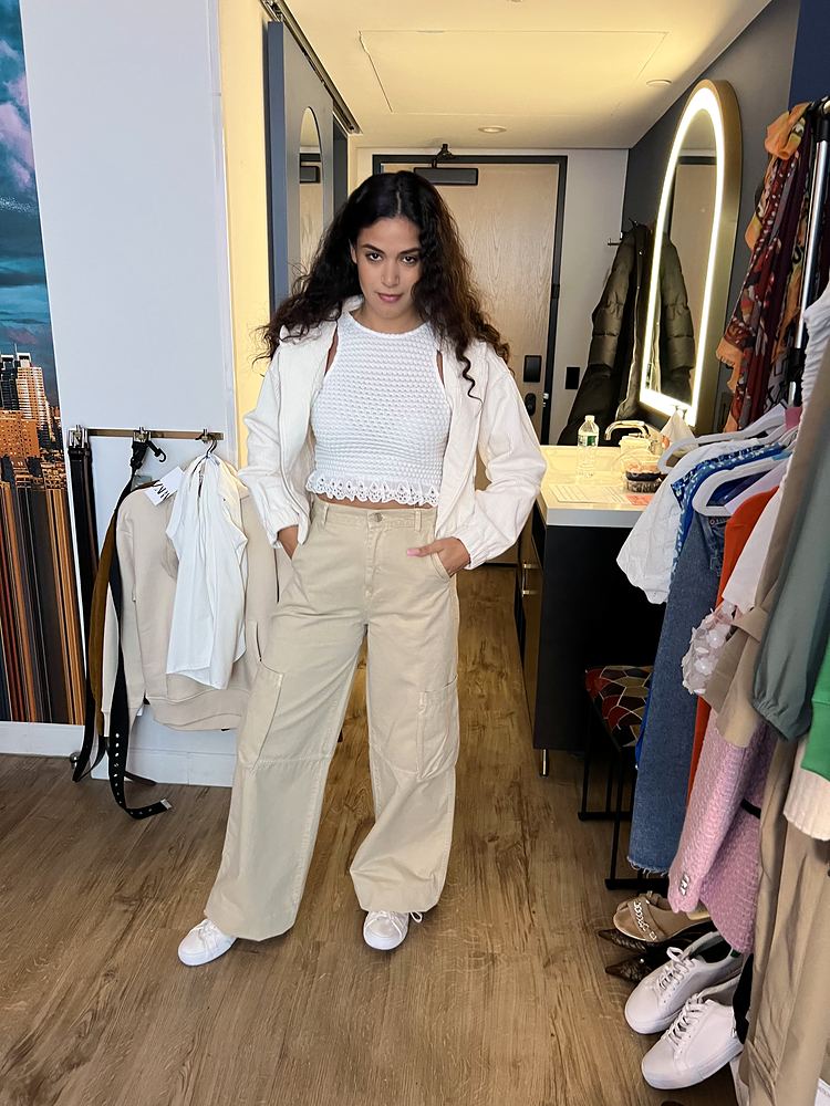 A woman with long curly hair in a white cardigan, white knit top and cream wide-leg trousers beside a wardrobe rack. Wardrobe design by Cynthia Nordstrom