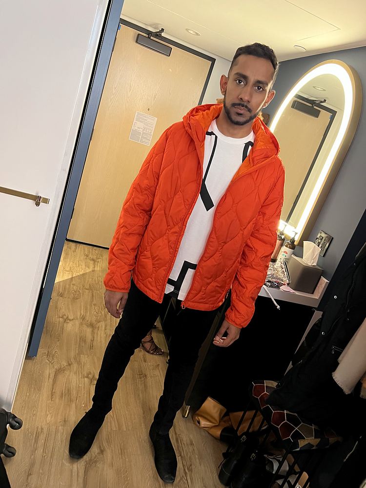 A man in an orange quilted puffer jacket, white T-shirt and black trousers in the hotel room used for fittings. Wardrobe design by Cynthia Nordstrom