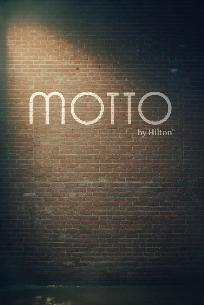 Motto by Hilton logo painted on a lamp-lit city brick wall, poster for the commercial