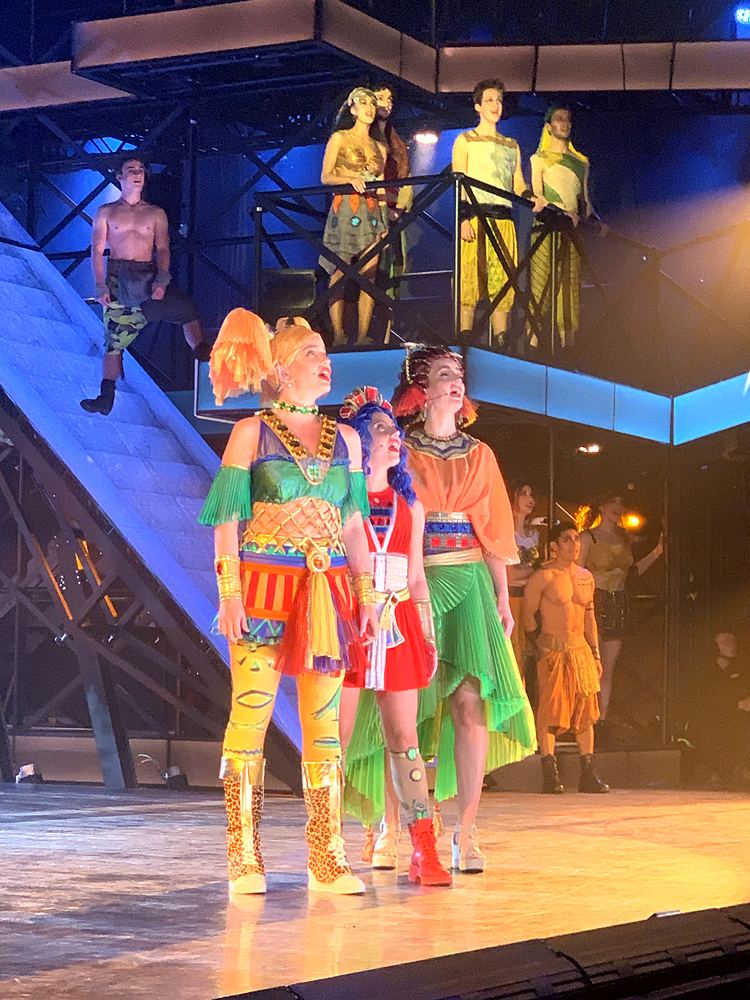 Women in bright patchwork costumes and tall boots on a multilevel set, ONE de Musical. Costume design by Cynthia Nordstrom