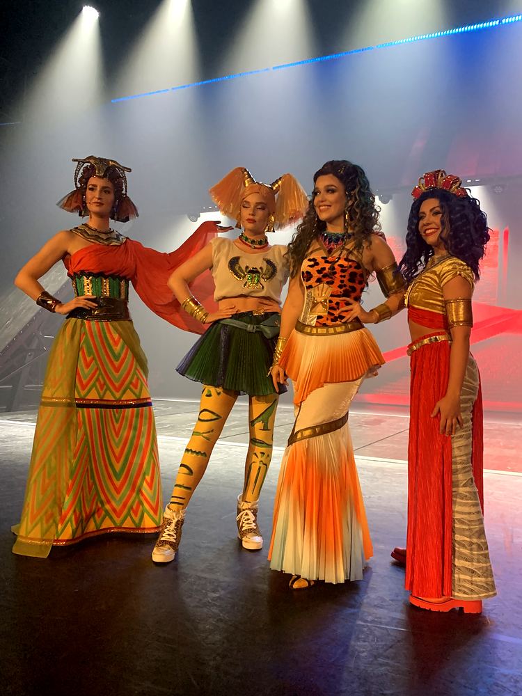 Four women in patterned gowns and headpieces lined up under spotlights, ONE de Musical. Costume design by Cynthia Nordstrom