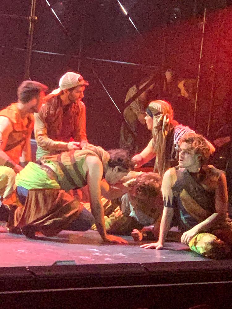 Young men in earthy rags and headwraps gathered on the floor, ONE de Musical. Costume design by Cynthia Nordstrom