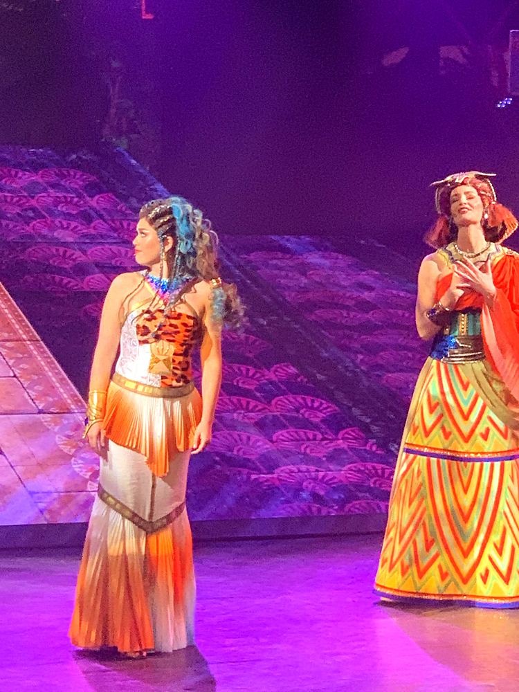 Two women in an orange and white gown and a yellow patterned gown, ONE de Musical. Costume design by Cynthia Nordstrom
