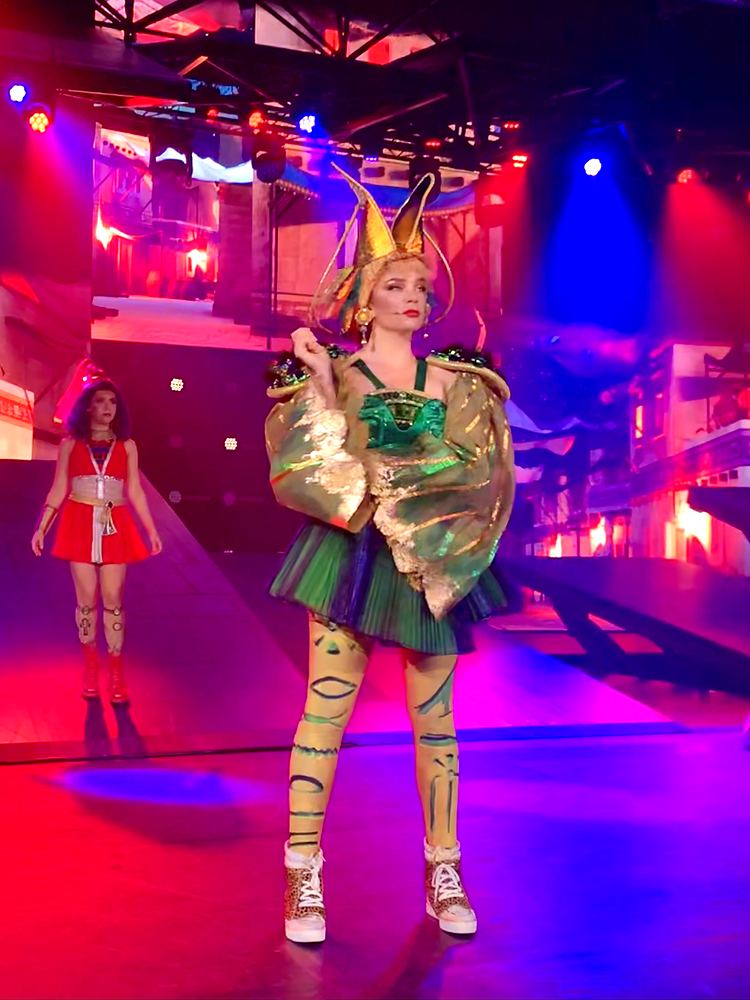Woman in a gold winged top, green skirt and painted tights under red light, ONE de Musical. Costume design by Cynthia Nordstrom