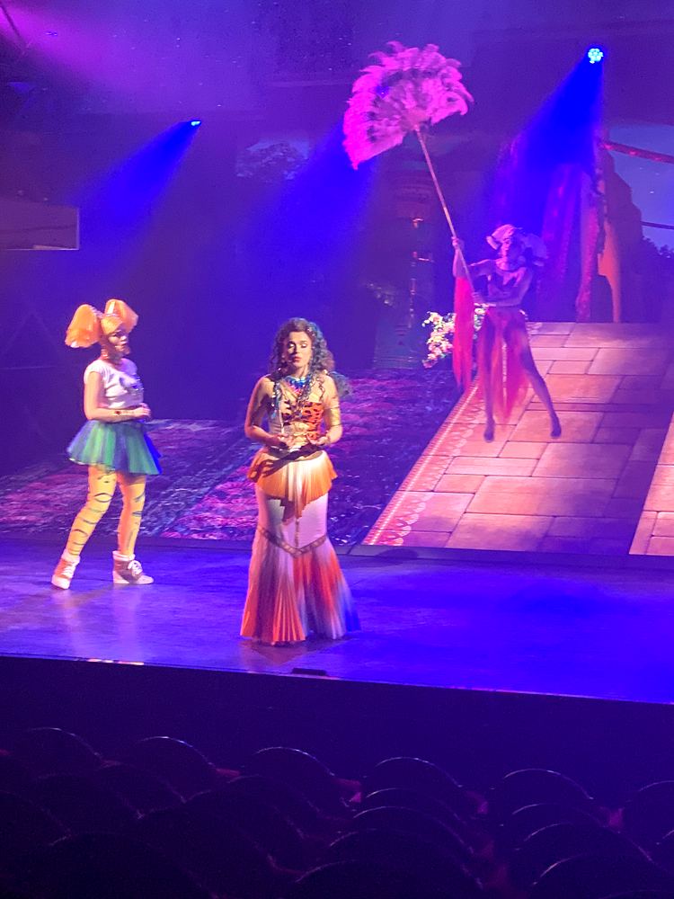 Two women in patterned skirts with a feather fan dancer behind, ONE de Musical. Costume design by Cynthia Nordstrom