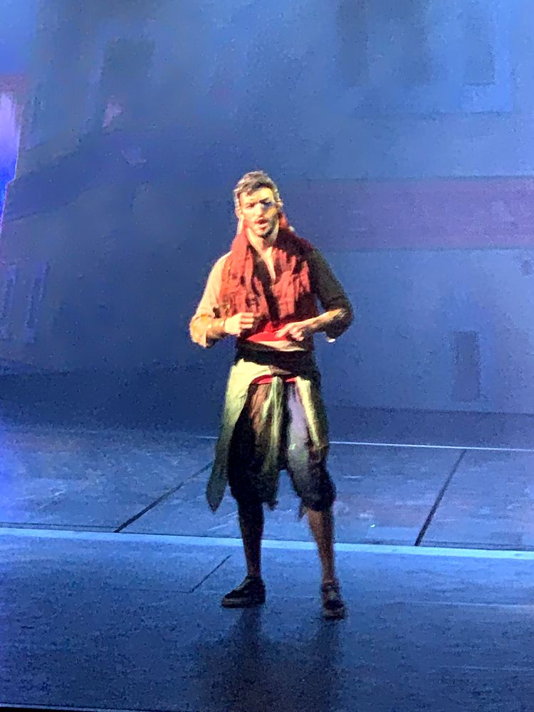 Man in a red scarf, black vest and wrapped sash alone on stage, ONE de Musical. Costume design by Cynthia Nordstrom