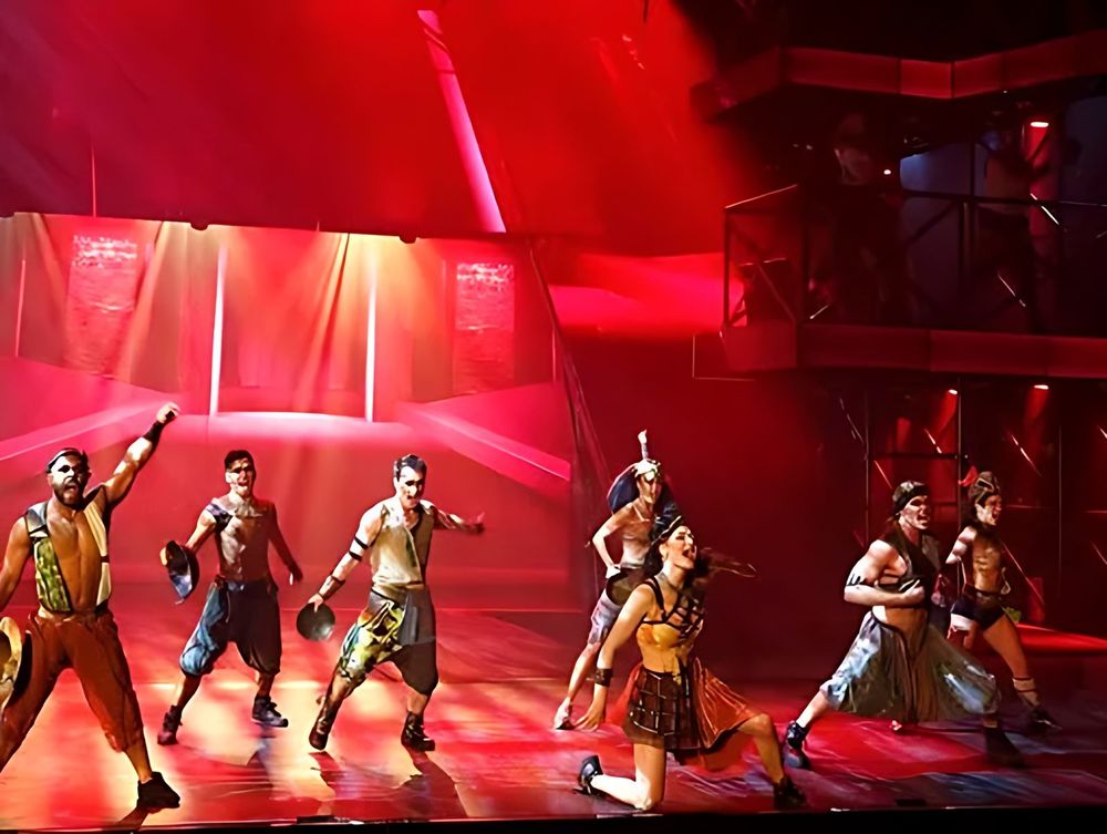 ONE de Musical: the ensemble on stage under red light. Costume design by Cynthia Nordstrom