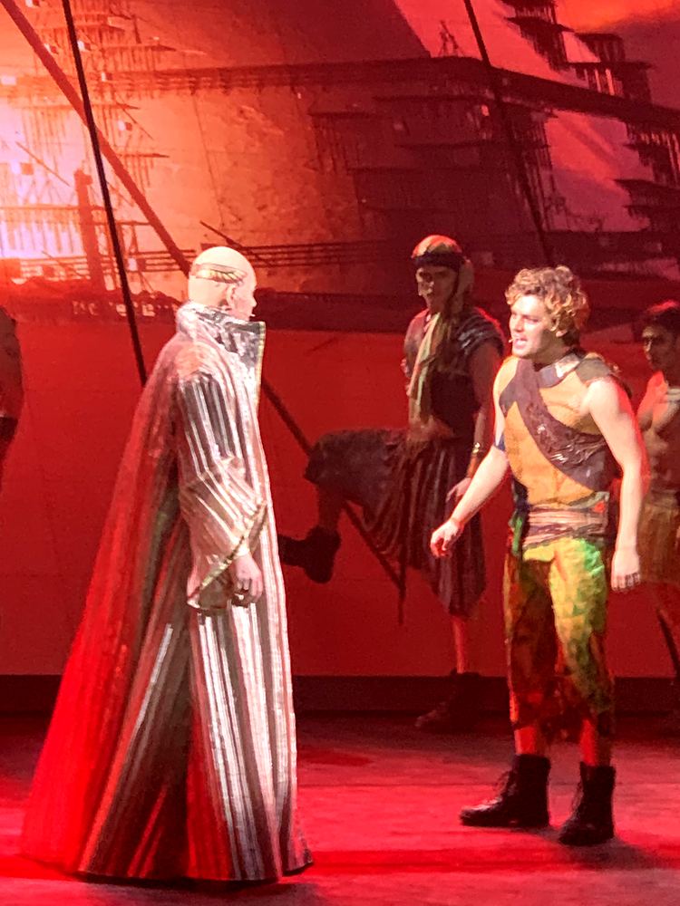 Elder in a striped cloak facing a young man in a leather harness, red light, ONE de Musical. Costume design by Cynthia Nordstrom