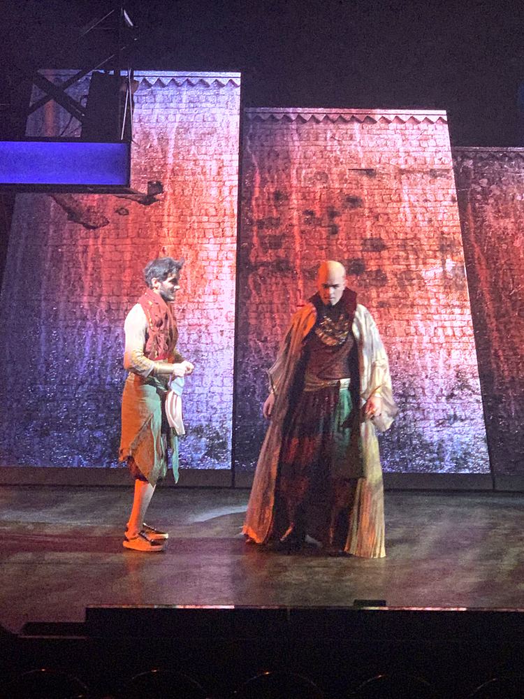 Two men in a patterned vest and long brown robe before tall stone walls, ONE de Musical. Costume design by Cynthia Nordstrom