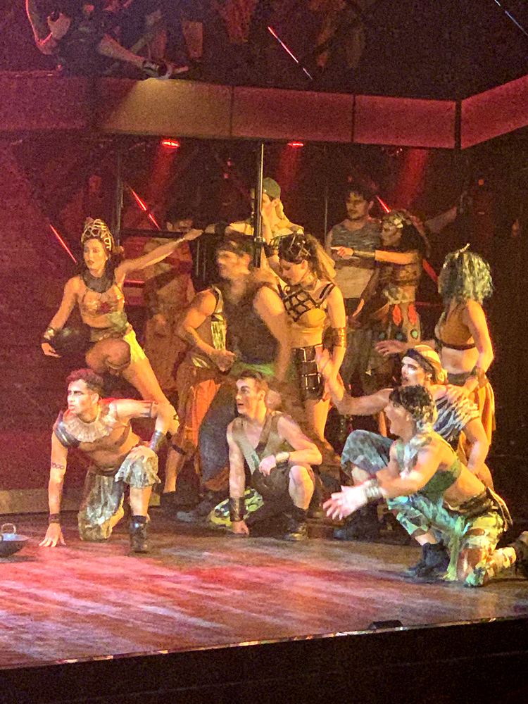 Ensemble in earth-tone warrior costumes posed in a group under red light, ONE de Musical. Costume design by Cynthia Nordstrom
