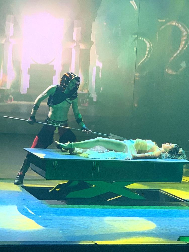 Masked figure with a spear over a woman lying on an altar in green light, ONE de Musical. Costume design by Cynthia Nordstrom