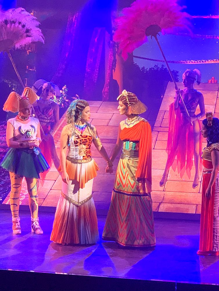 Women in patterned gowns and headdresses with feather fans behind, ONE de Musical. Costume design by Cynthia Nordstrom