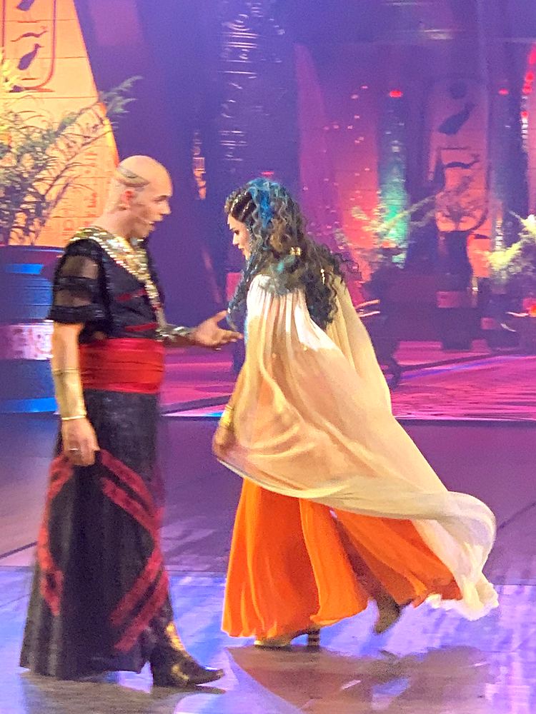 Woman in a cream cape and orange skirt approaching a bald man in black and red, ONE de Musical. Costume design by Cynthia Nordstrom