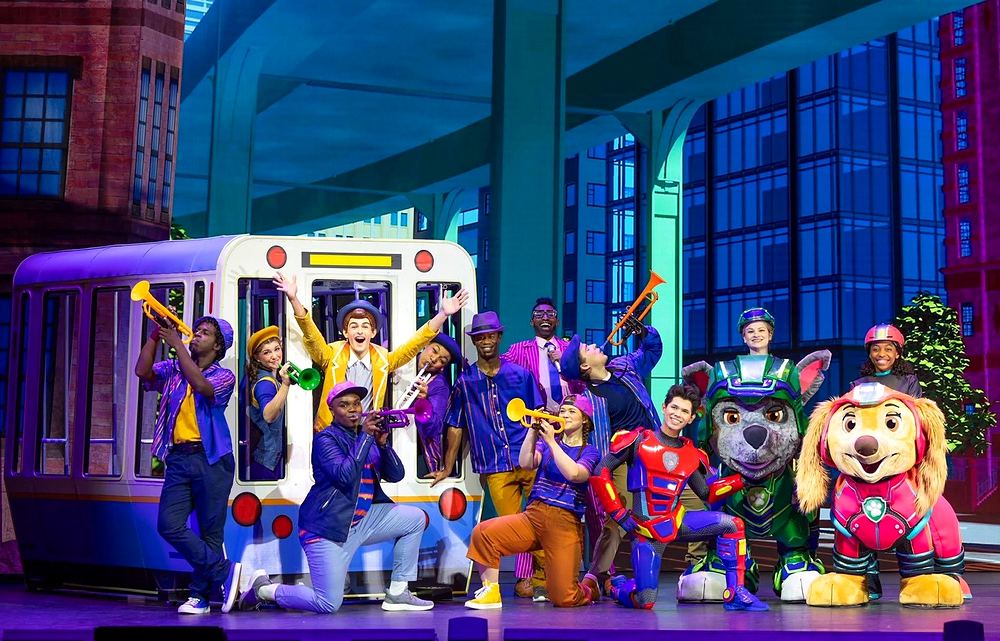 PAW Patrol Live! A Mighty Adventure: the company in purple, blue and striped band uniforms with trumpets around a city tram, with Ryder, Rocky and Skye. Costume design by Cynthia Nordstrom
