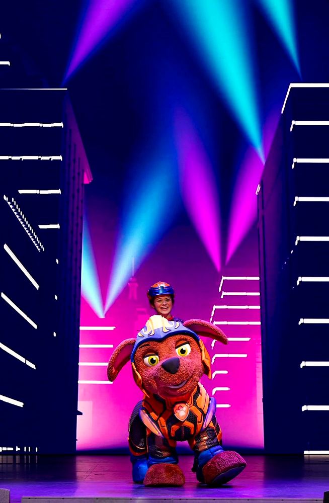 PAW Patrol Live! A Mighty Adventure: Zuma in orange and navy Mighty Pups armor with his helmeted handler under pink and teal light. Costume design by Cynthia Nordstrom