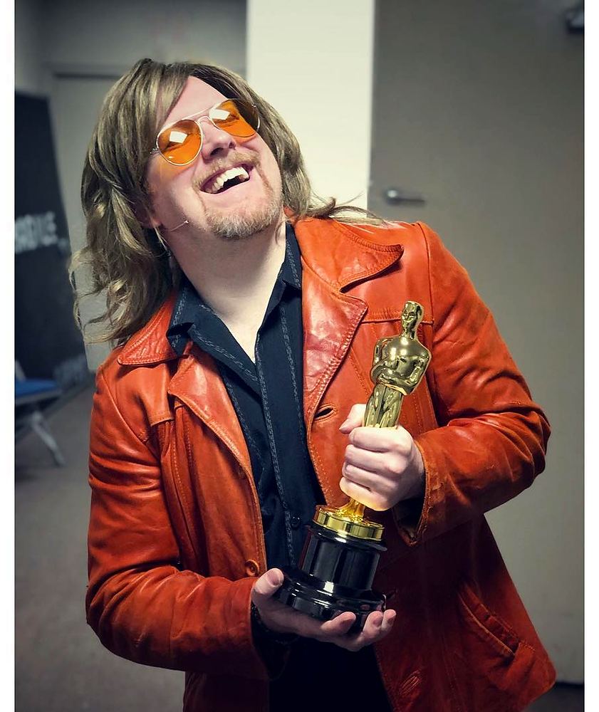Grinning rocker in an orange leather jacket and tinted glasses with a trophy, Rock of Ages. Costume design by Cynthia Nordstrom