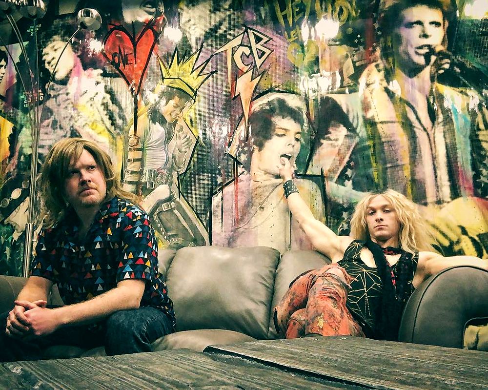 Two rockers in a patterned shirt and leather lounging before a rock mural, Rock of Ages. Costume design by Cynthia Nordstrom