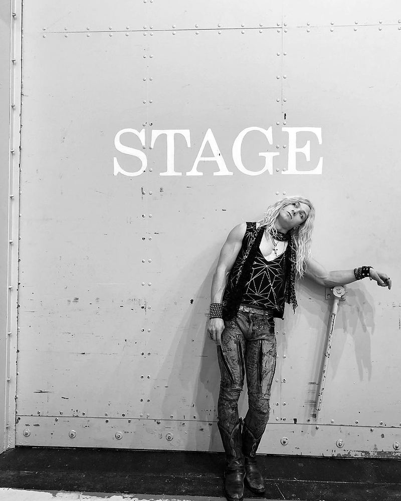 Rocker in a studded vest and leather pants under a STAGE sign, black and white, Rock of Ages. Costume design by Cynthia Nordstrom