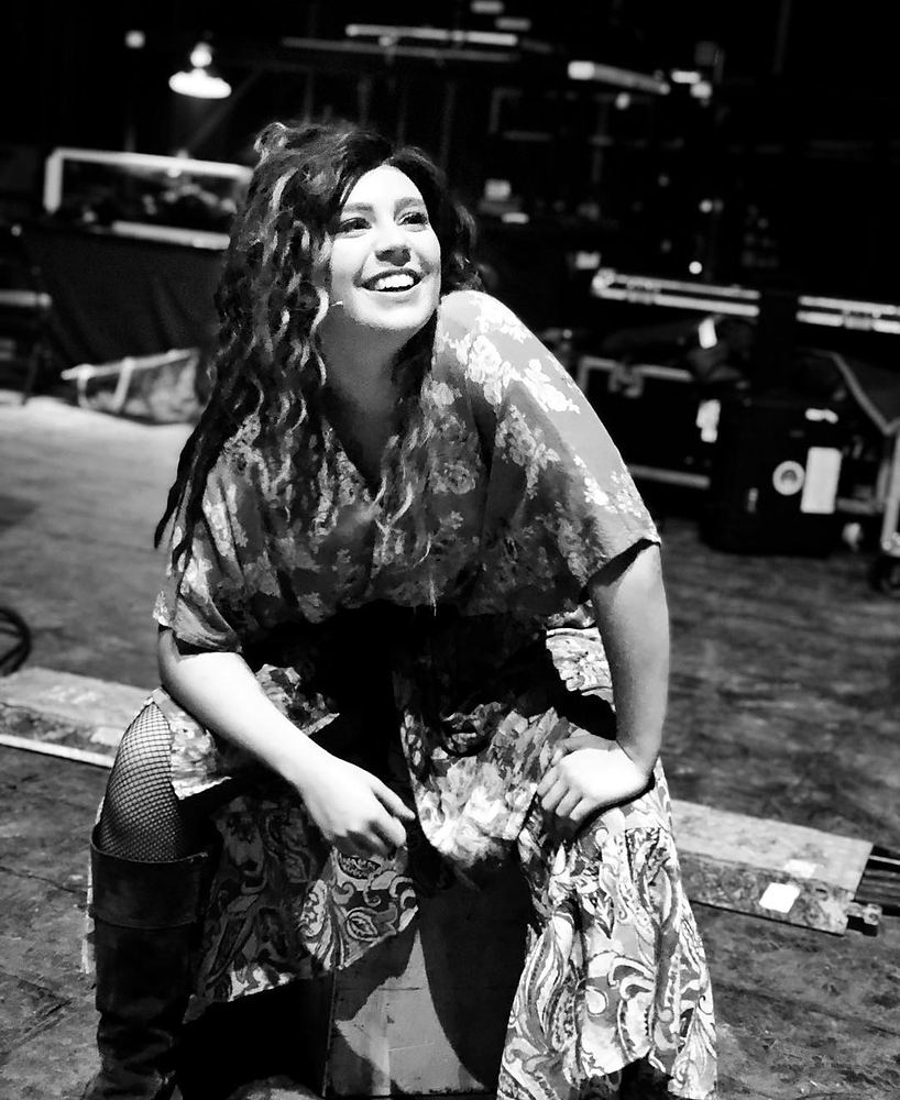 Smiling woman in a print caftan and boots sitting backstage, Rock of Ages. Costume design by Cynthia Nordstrom