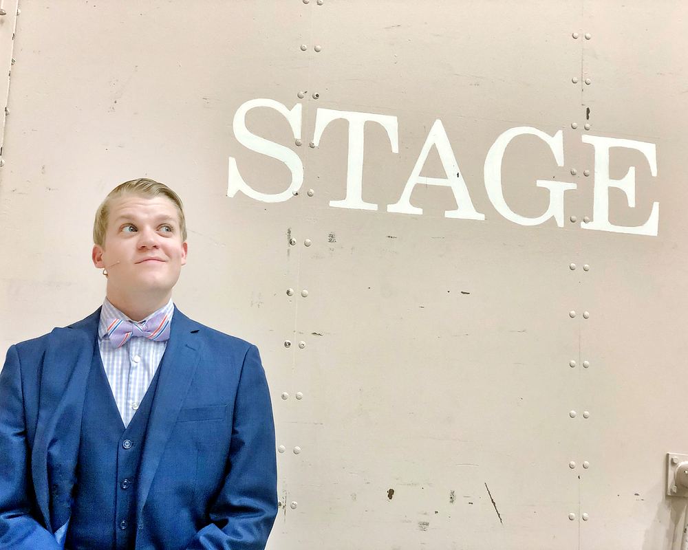 Young man in a blue three-piece suit and bow tie under a STAGE sign, Rock of Ages. Costume design by Cynthia Nordstrom