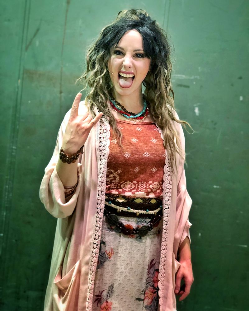 Woman in a pink duster, red top and layered skirt making a rock sign, Rock of Ages. Costume design by Cynthia Nordstrom