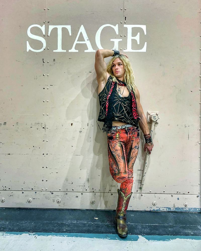 Rocker in a studded vest and red patterned pants with a sword under a STAGE sign, Rock of Ages. Costume design by Cynthia Nordstrom