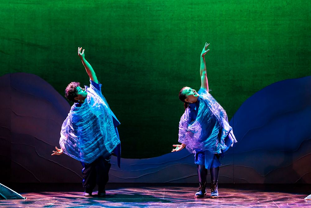 Two dancers in flowing blue and purple capes against a green sky, Spells of the Sea. Costume design by Cynthia Nordstrom