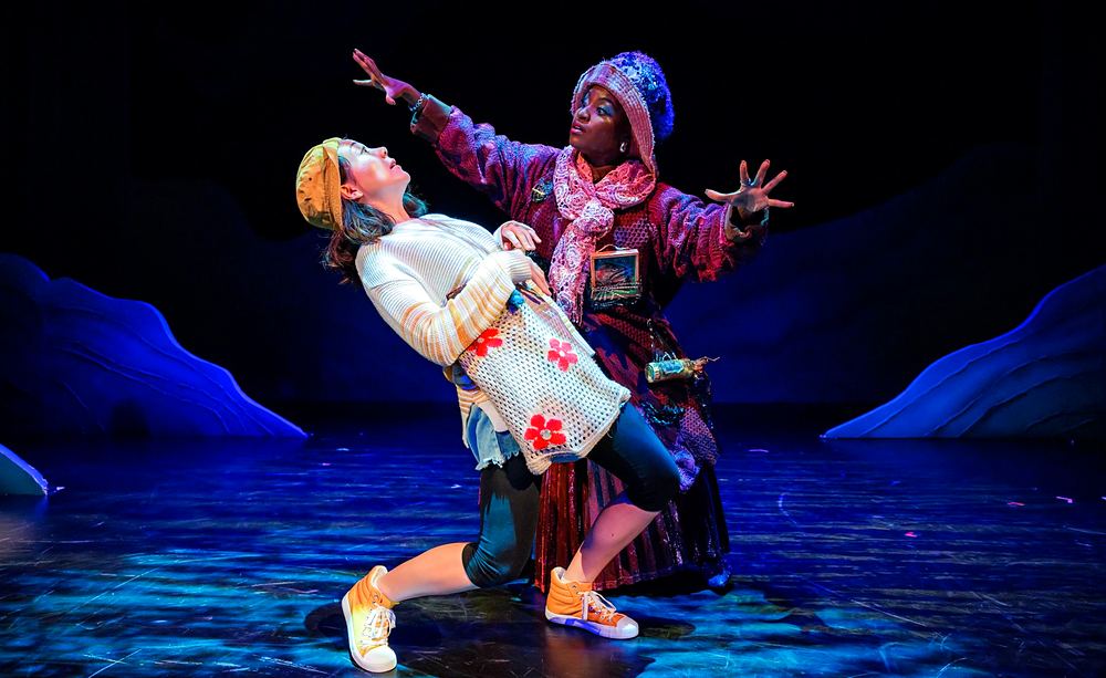 Shopkeeper in a red patchwork coat looming over a girl in a floral top, Spells of the Sea. Costume design by Cynthia Nordstrom