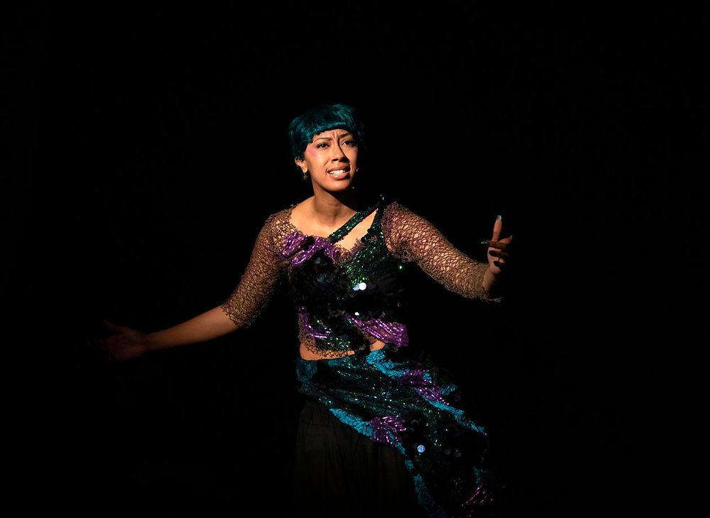 Performer in a sparkling teal and black sea witch costume in a spotlight, Spells of the Sea. Costume design by Cynthia Nordstrom
