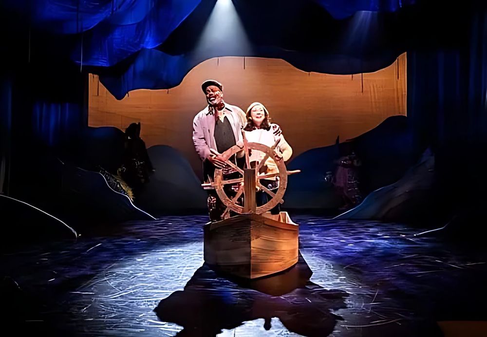 Captain and girl at a ship wheel on a small boat under a spotlight, Spells of the Sea. Costume design by Cynthia Nordstrom