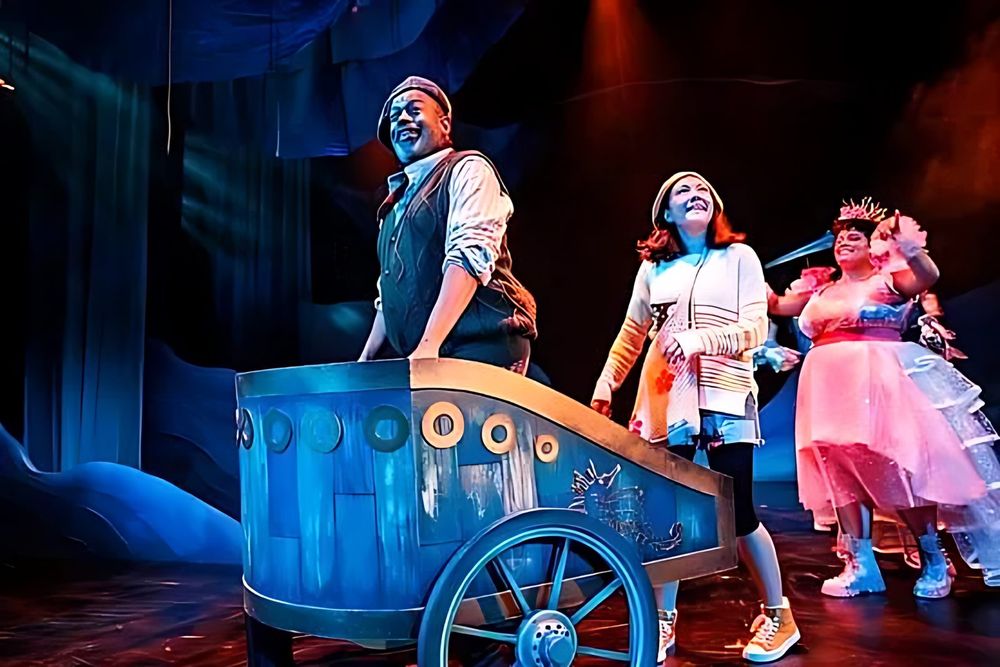 Man in a vest pushing a blue cart as girls in patterned layers follow, Spells of the Sea. Costume design by Cynthia Nordstrom
