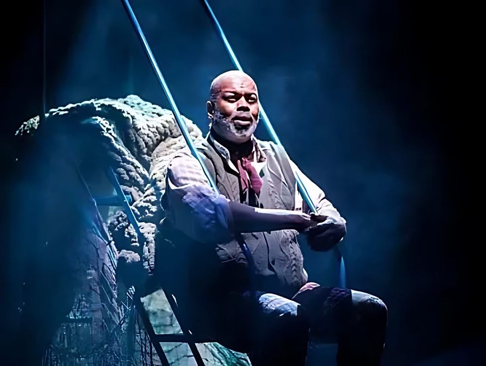 Older man in a vest and scarf sitting on a rock in misty light, Spells of the Sea. Costume design by Cynthia Nordstrom