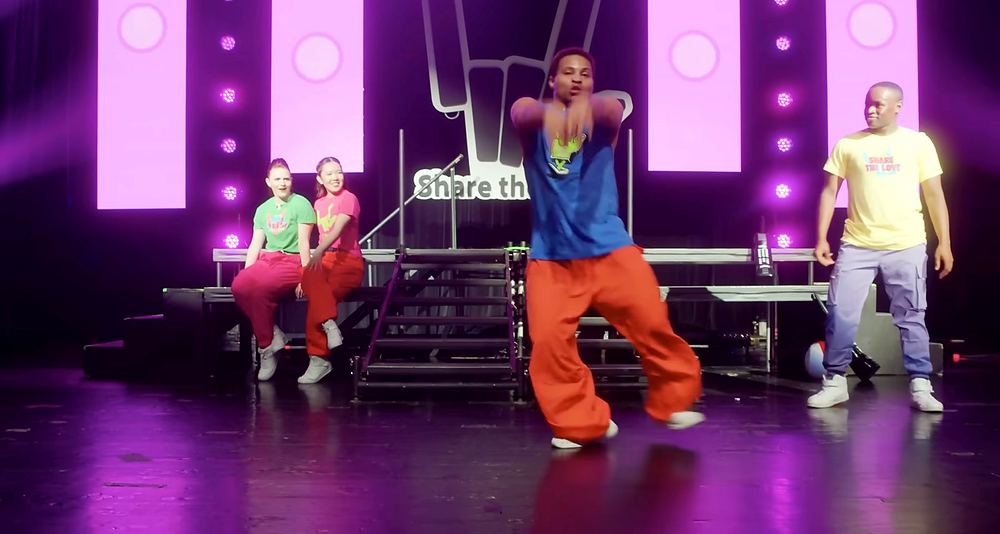 Dancers in neon tees and bright pants on a pink stage with a hand logo screen, Stephen Sharer: Share the Love Tour. Co-costume design by Cynthia Nordstrom