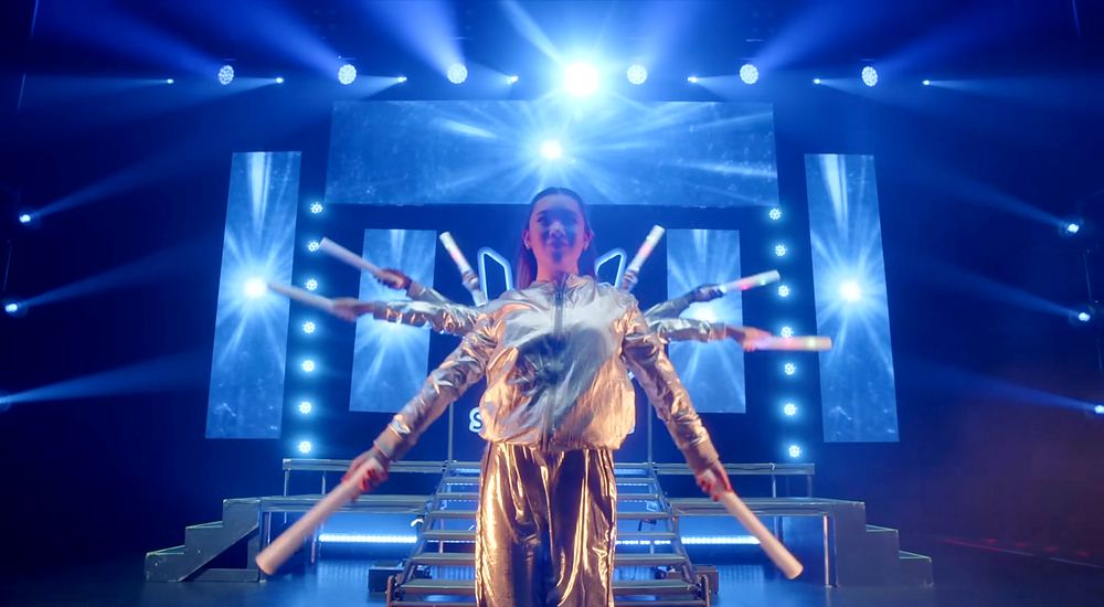 A dancer in a brown feathered costume on a blue lit stage with star shaped lights, Stephen Sharer: Share the Love Tour. Co-costume design by Cynthia Nordstrom