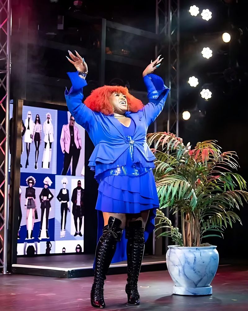 Woman in a red wig, blue ruffled jacket and skirt and tall black boots, SuperYou. Costume design by Cynthia Nordstrom