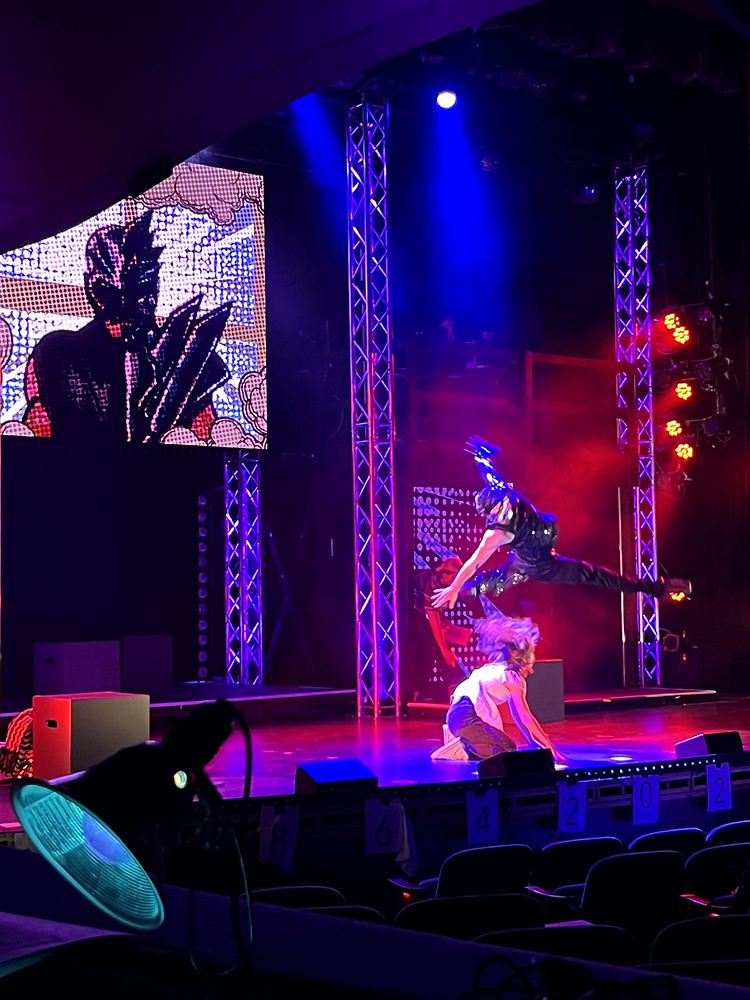 Hero in black leaping across the stage under blue and red lights, SuperYou. Costume design by Cynthia Nordstrom