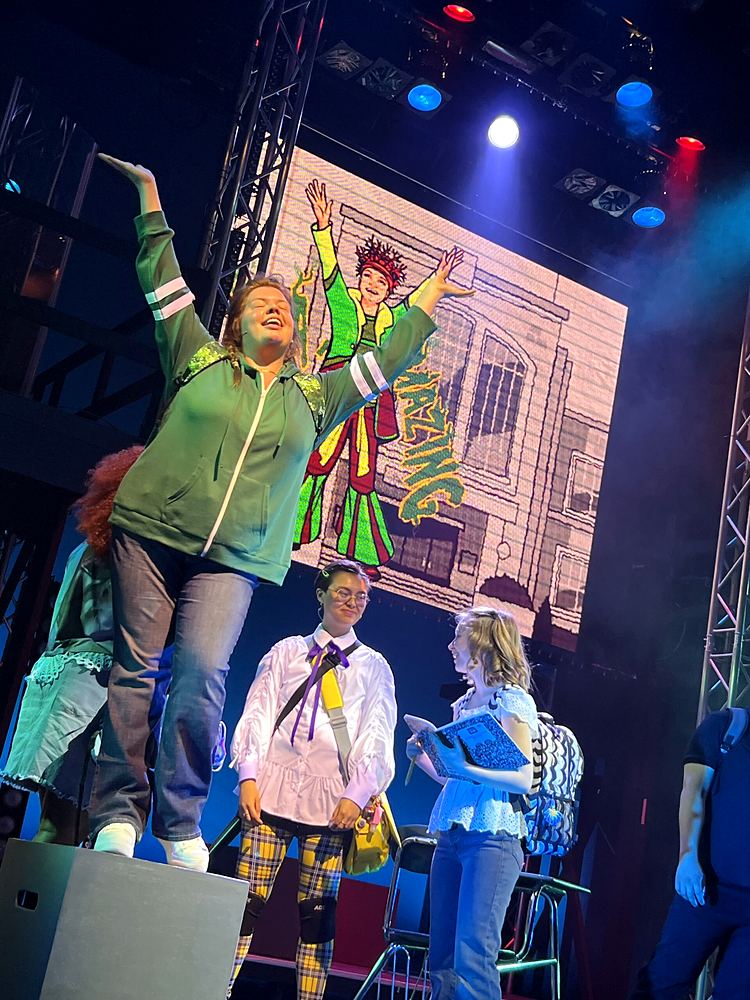 Woman in a green hoodie raising her arms with the cast on a comic book set, SuperYou. Costume design by Cynthia Nordstrom