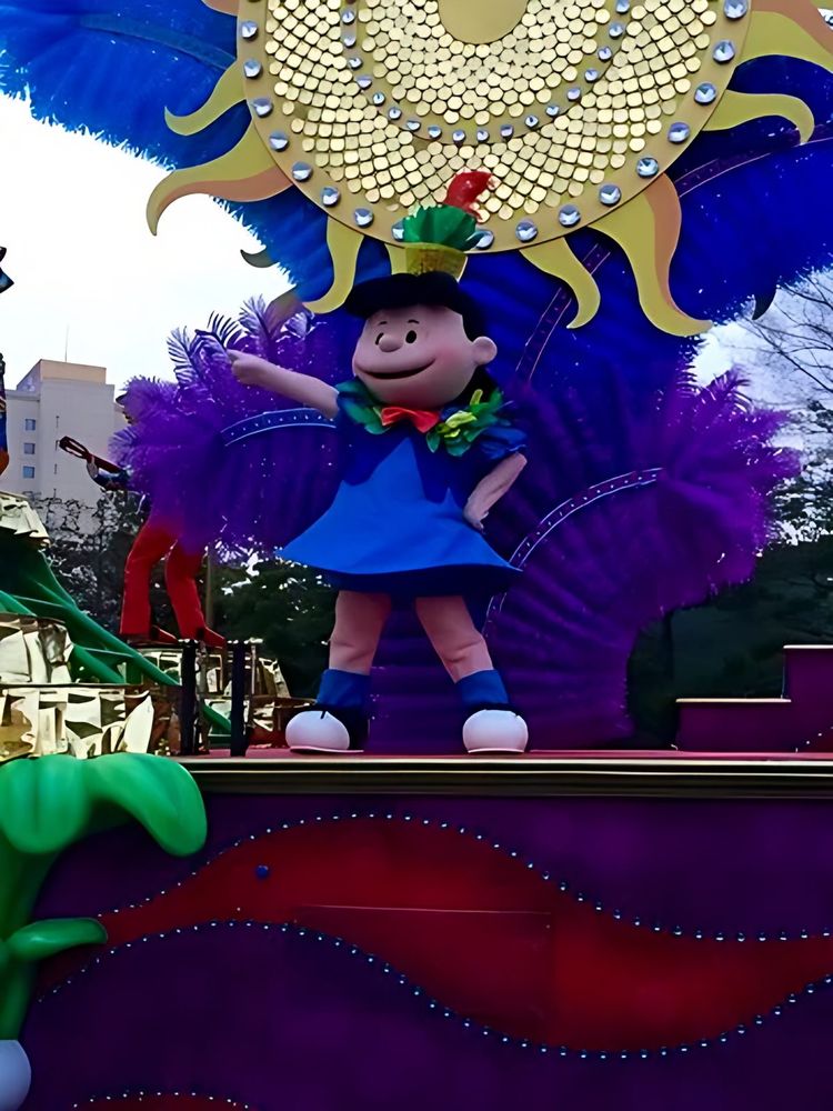 Lucy in a blue dress waving from a float with a sun and purple feathers, USJ anniversary shows. Costume design by Cynthia Nordstrom