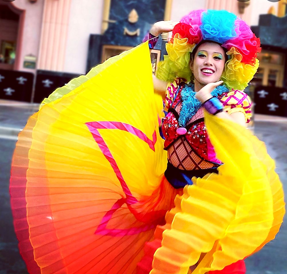 Smiling dancer in a rainbow wig spreading a yellow and orange pleated skirt, Universal Beijing parade. Costume design by Cynthia Nordstrom